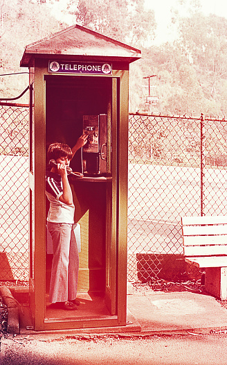 Vintage image of a boy talking by phone in a public telephone booth of the seventies/eighties of the 20th century.