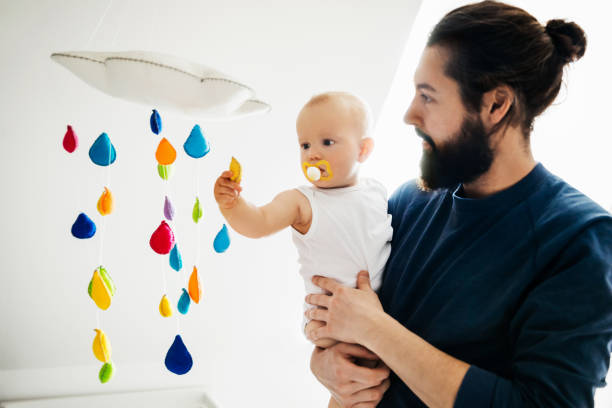 Baby Holding Onto Dad While Playing Wth Mobile A single dad holding onto his baby while he plays with a colourful mobile. hanging mobile stock pictures, royalty-free photos & images