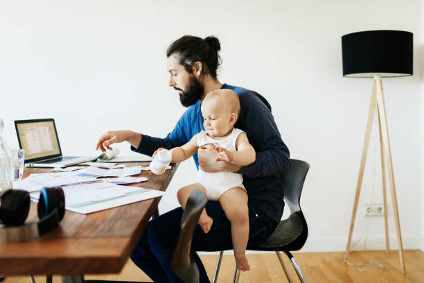 Single Father Working While Holding Baby A single father working at his desk at home while holding his baby son. working at home with children stock pictures, royalty-free photos & images