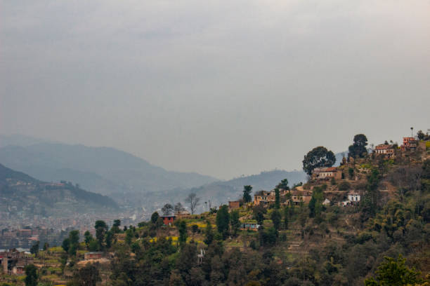 A wide-view shot of a town located on the side of a large hill, the town is surrounded by nature with trees growing all around the houses. The image is on a foggy day.