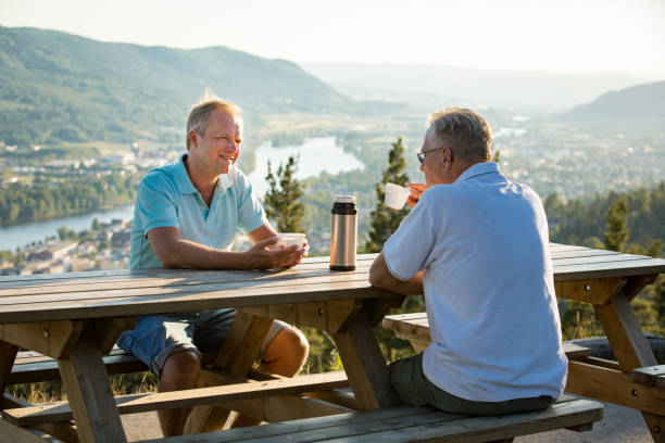 Two mature men sitting at wooden table on observation deck on top of mountain Two mature men sitting at wooden table on observation deck on top of mountain, drink hot coffee from thermos flask talking, smiling, laughing. Beautiful view of city in valley in haze. Norway, Drammen østfold stock pictures, royalty-free photos & images