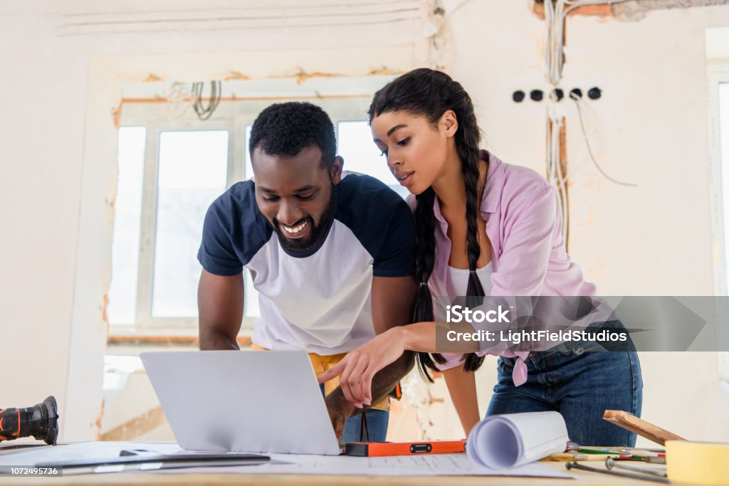 beautiful african american woman pointing at laptop screen to smiling boyfreind during renovation of home Home Improvement Stock Photo