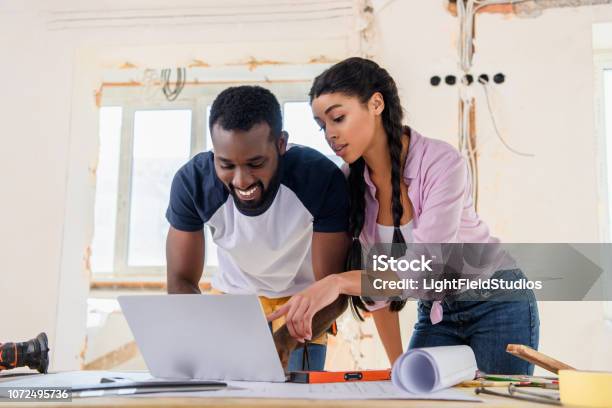 Beautiful African American Woman Pointing At Laptop Screen To Smiling Boyfreind During Renovation Of Home Stock Photo - Download Image Now