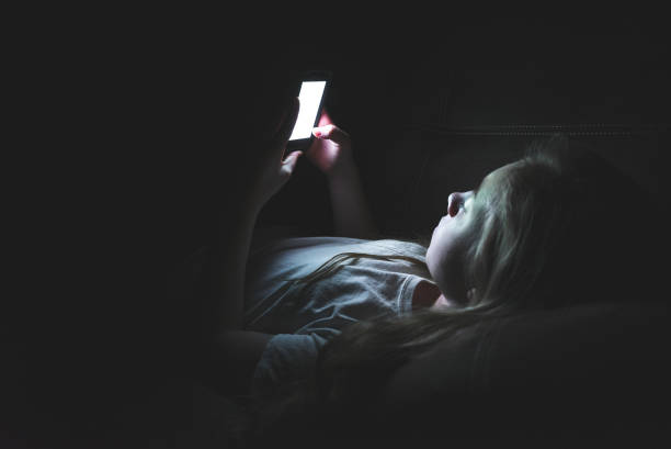 Depressed girl on her smartphone while lying down in the dark. stock photo