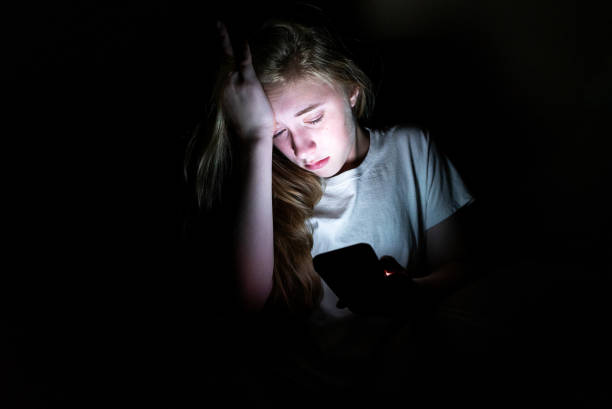 Upset girl on her smartphone in the dark. The image displays an upset girl sitting in the dark while using her smartphone. The light from the screen is illuminating her face. bullying photos stock pictures, royalty-free photos & images