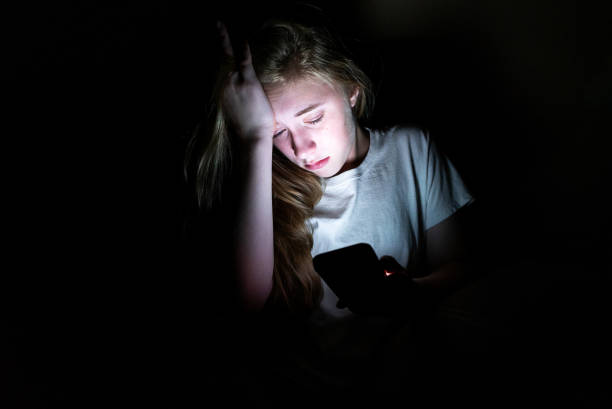 Upset girl on her smartphone in the dark. The image displays an upset girl sitting in the dark while using her smartphone. The light from the screen is illuminating her face. child sadness stock pictures, royalty-free photos & images