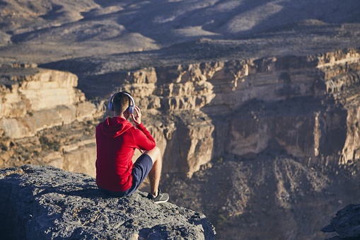 Relaxation in mountains. Young man with headphones sitting on the edge of cliff and listening music. Jebel Akhdar, Grand Canyon of Oman.