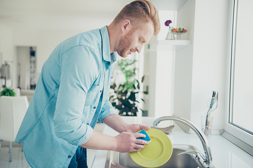 Good-looking man in blue shirt casual stylish trendy wear washing green dishes carefully and attentively stand inside bright flat