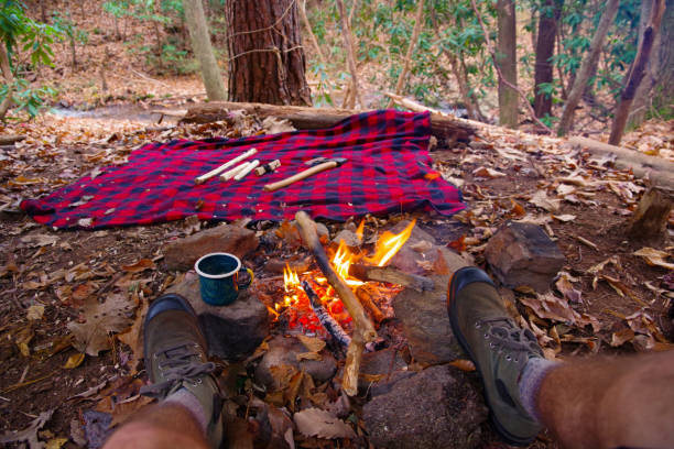 First person view of man relaxing / having tea in an enamel outdoor mug by a campfire in Blue Ridge / Appalachian Mountains trail near Asheville, North Carolina. Boots, Bushcraft, blanket, Axe. stock photo