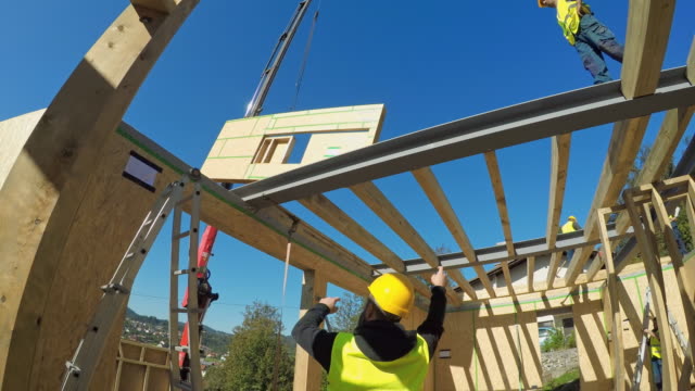 Wide handheld shot of a male foreman signaling the worker operating the crane to move a wooden wall for the prefab home to the upper level of the house. Shot in Slovenia.