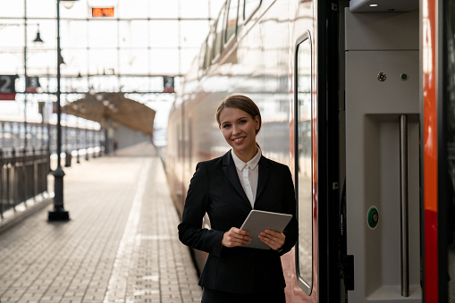 Portrait of a beautiful train attendant smiling at the platform and looking at the camera holding a tablet computer - rail transportation concepts