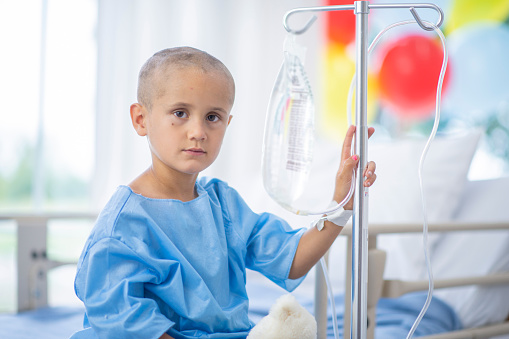 A young boy with a shaved head is sitting on a hospital bed. The boy has cancer, and he is attached to an IV. He is staring at the camera.