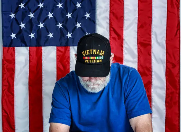 Authentic 68 year old United States Navy Vietnam War military veteran sitting looking down with his face partially covered. Copy space on the USA flag stars and stripes red, white and blue background. He is wearing an inexpensive, non-branded, generic, souvenir shop replica Vietnam veteran commemorative baseball hat style cap.
