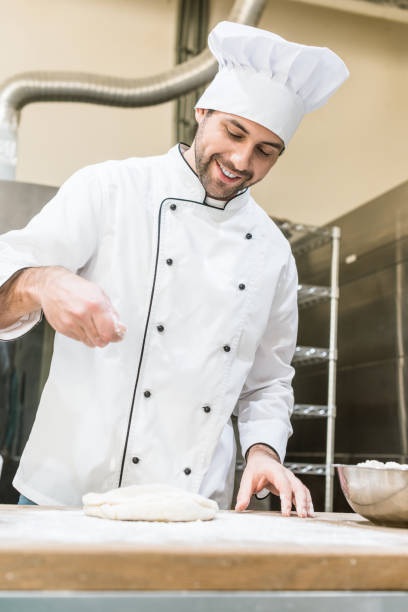 istock baker in white chefs uniform smiling and cooking dough in professional kitchen 1072630560