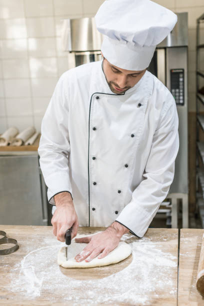 istock Baker in white chefs uniform cutting dough on wooden table at professional kitchen 1072630606