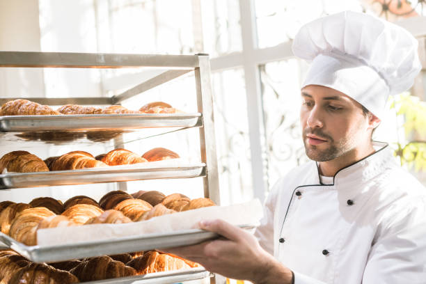 istock baker in chefs uniform looking at trays with fresh cooked croissants 1072635548