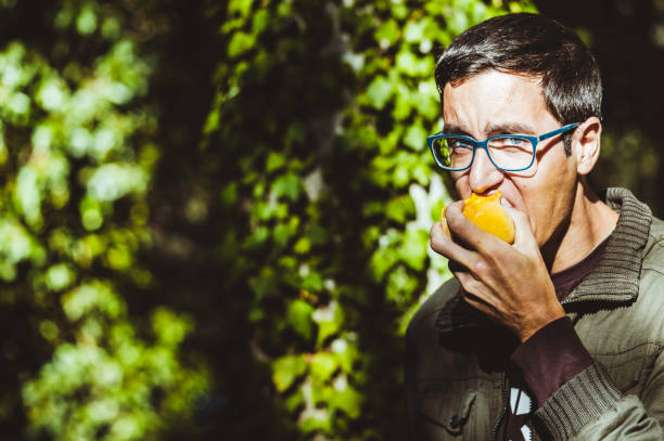 istock Young man with glasses and blue eyes biting a fruit, peach, with green leaves 1072635820