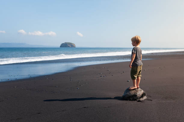 Little kid stand on big stone on black sand sea beach. Dreaming child look at sea surf, waves. Solitude concept. Retreat leisure on summer family vacation