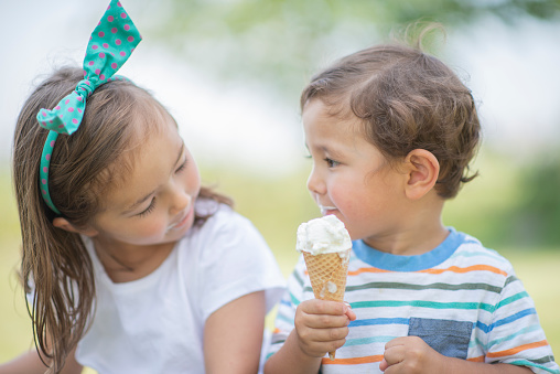 A boy and girl are outdoors in a park on a summer day. They are sharing an ice cream cone.