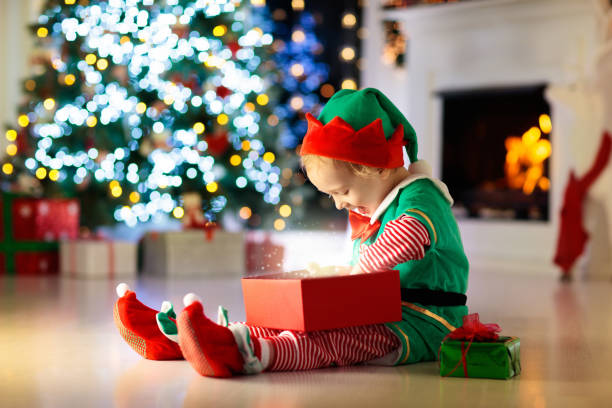Child opening present at Christmas tree at home. Kid in elf costume with Xmas gifts and toys. Little baby boy with gift box and candy at fireplace. Family celebrating winter holidays. Home decoration.
