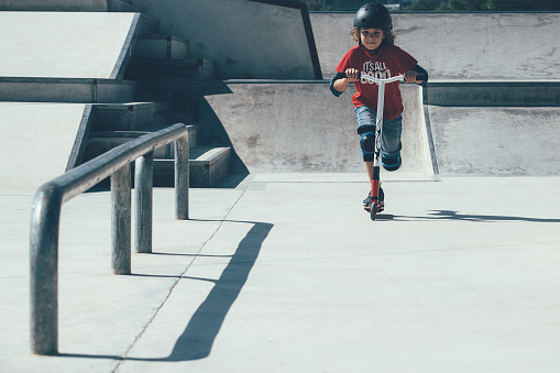 Front view of a young boy wearing a helmet and the protections is using a scooter in an urban skate park.