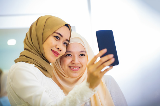 Young Muslim woman taking a selfie with her friend who is trying on a wedding dress in the bridal shop's fitting room.