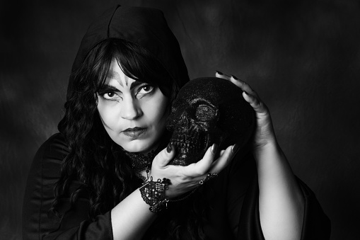 Horizontal black and white studio shot on dark background of mature Native Canadian woman with sorceress makeup holding black skull.  Head and shoulders.