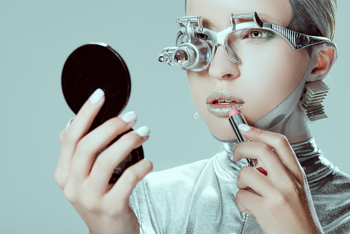 silver robot looking at mirror and applying lipstick isolated on grey, future technology concept