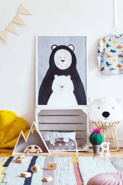 istock Stylish scandinavian kid room with mock up photo poster frame on the pattern wall, boxes, teddy bear and toys. Cute modern interior of playroom with white walls, wooden accessories and colorful toys. 1072751064