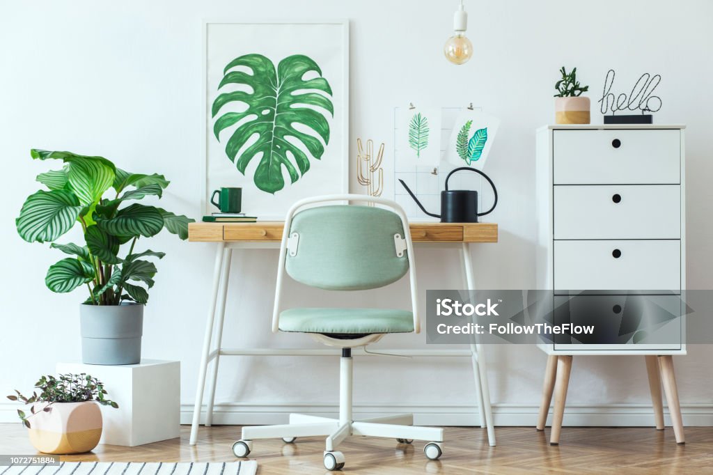 Stylish scandinavian home office desk with mock up poster frames, a lot of plants and office accessories. Brown wooden parquet and white backgrounds wall. Modern composition of homeoffice desk. Stylish scandinavian home office desk with mock up poster frames. Home Office Stock Photo