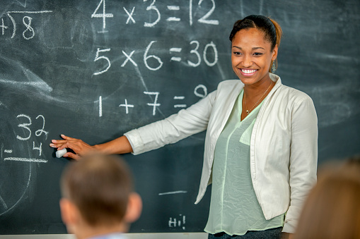 A black female teacher is giving a math lesson in a classroom. She is standing in front of a chalkboard that has math equations on it.