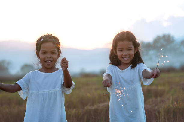 two cute asian child girl are playing with fire sparklers on the festival in the rice field - happy kid flash imagens e fotografias de stock