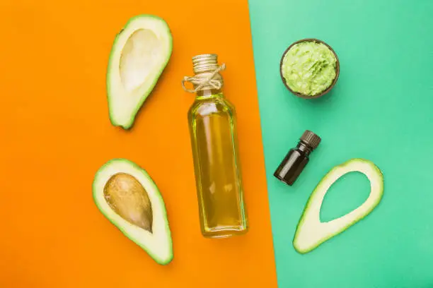 Bottle natural avocado oil and essential oil for healthy face and body. Cosmetic mask for healthy skin and beautiful hair. Fresh tropical fruit avocado cut in half lies on an orange and green table. Top view