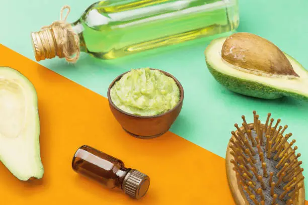 Mask avocado for healthy and beautiful hair in wooden bowl, hairbrush, olive oil and essential oil in a bottle. Fresh fruit of avocado on a color background, orange and green.
