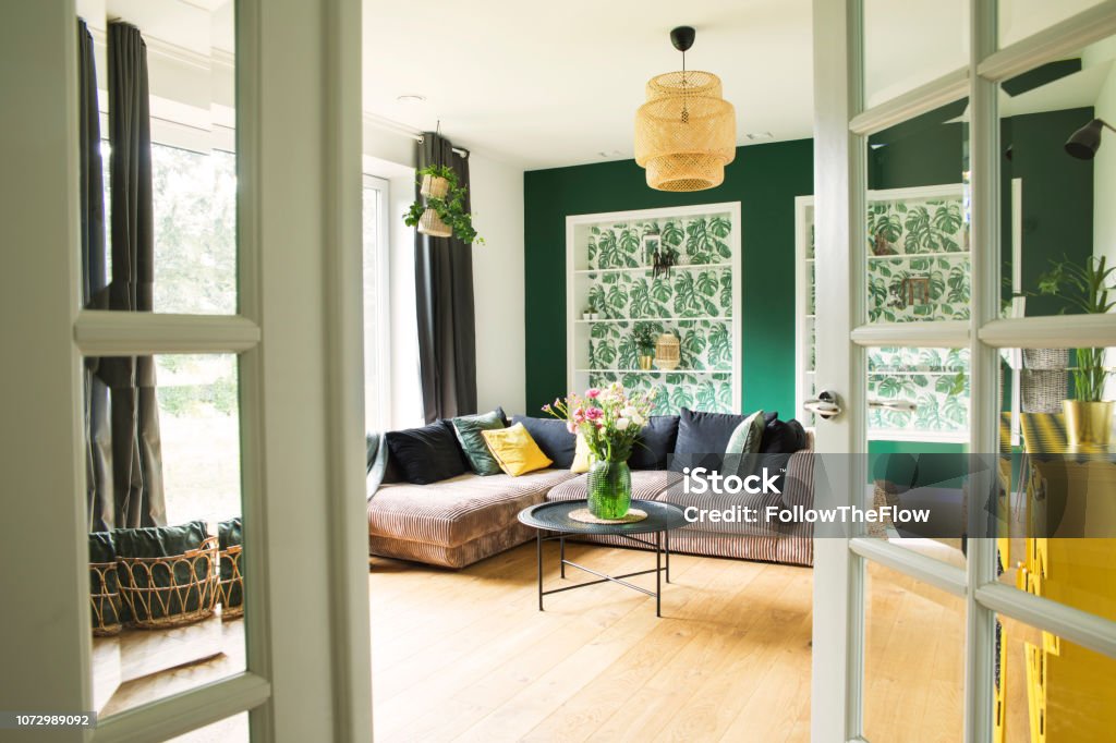 Stylish interior with sofa, colors pillows, flowers and wooden floor. Sunny and bright living room with big window and design commode.. Stylish and cozy living room- modern interior composition. Green Color Stock Photo