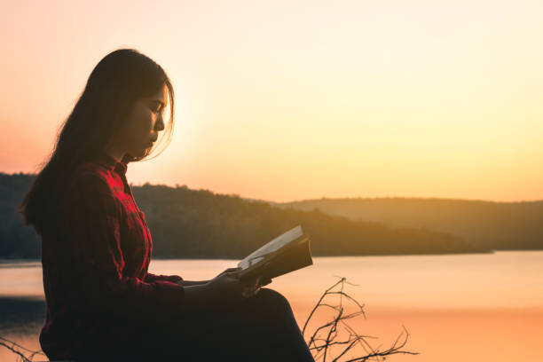 istock Woman Reading Book While Sitting By Lake During Sunset 1072996560