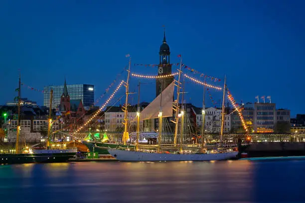 A festively decorated sailing ship with 4 masts at blue hour at the pier in the port of Hamburg in front of the Michel - the landmark of the city