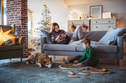 Young mother with son celebrating Christmas at home. Home is decorated with Christmas ornaments and lights. Boy plays with dog on floor