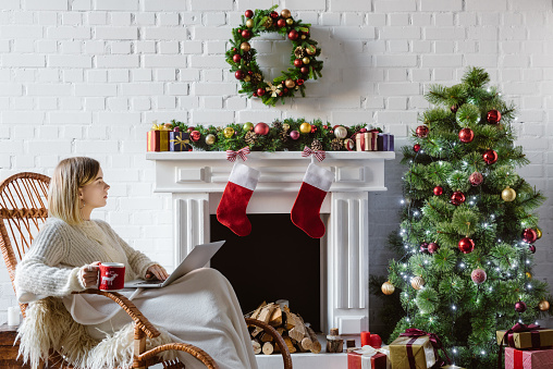 beautiful young woman on wicker rocking chair holding coffee cup and using laptop in living room decorated for christmas