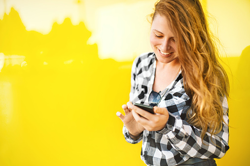 Young beautiful woman checking new amazing app on her smartphone. She's standing in front of a yellow wall. Shes excited.
