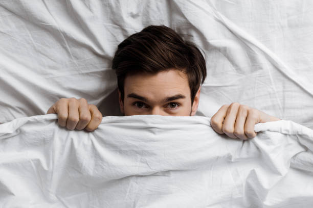 istock top view of young man hiding under blanket in bed and looking at camera 1073105712