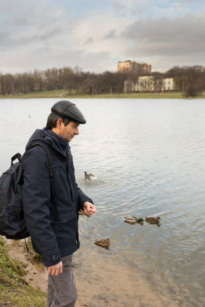 istock Handsome dark-haired middle-aged man in jacket, cap and backpack feeds ducks on pond in city park in late autumn. Authentic lifestyle moments 1073124132