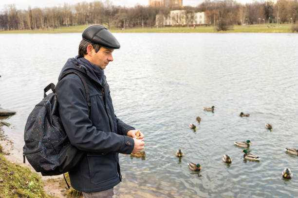istock Handsome dark-haired middle-aged man in jacket, cap and backpack feeds ducks on pond in city park in late autumn. Authentic lifestyle moments 1073124190