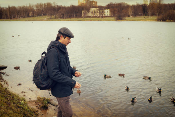 istock Handsome dark-haired middle-aged man in jacket, cap and backpack feeds ducks on pond in city park in late autumn. Authentic lifestyle moments 1073124250