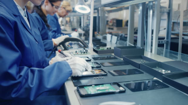 Female Electronics Factory Worker in Blue Work Coat and Protective Glasses is Assembling Smartphones with Tweezers and Screwdriver. High Tech Factory Facility with more Employees in the Background.