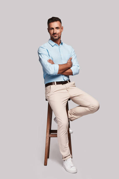 Totally handsome. Full length of good looking young man smiling and keeping arms crossed while sitting on stool against grey background business casual fashion stock pictures, royalty-free photos & images
