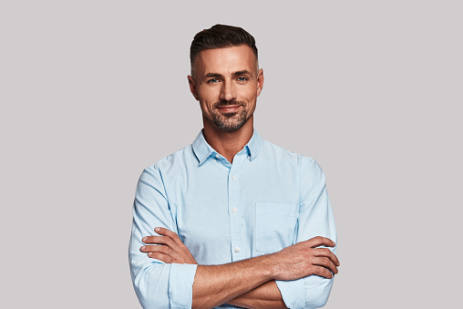 Charming young man looking at camera and smiling while standing against grey background