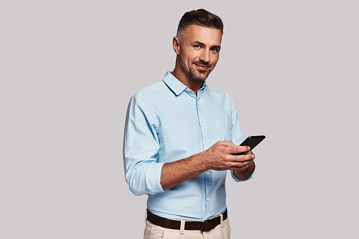 Good looking young man in smart casual wear using his smart phone and smiling while standing against grey background