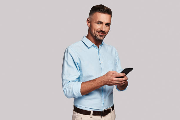 Always available. Good looking young man in smart casual wear using his smart phone and smiling while standing against grey background business casual fashion stock pictures, royalty-free photos & images