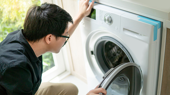 Young Asian technician looking into washing machine checking inside. Appliance maintenance in laundry at home. Technology concept